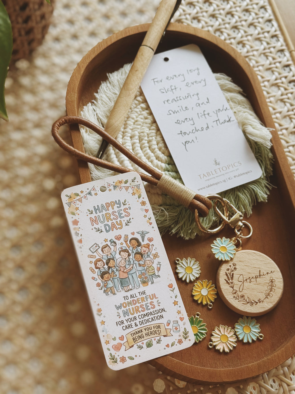 A wooden tray holds a personalized nurse-themed gift tag with a floral illustration and message, a white hanging tag with handwritten text, a tan pen, a green fringed cord, and small daisy charms on a woven surface.