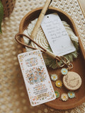 A wooden tray holds a personalized nurse-themed gift tag with a floral illustration and message, a white hanging tag with handwritten text, a tan pen, a green fringed cord, and small daisy charms on a woven surface.