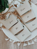 A collection of beige-colored drink sleeves with names written on them, displayed on a table with a decorative white and beige runner.